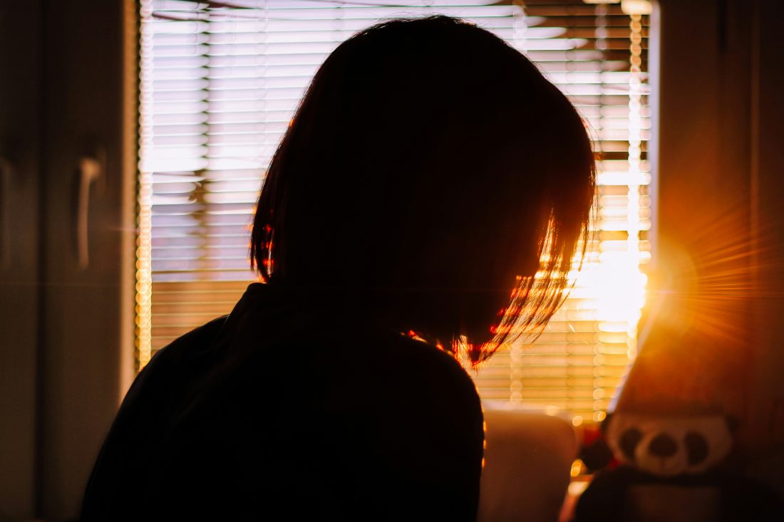 Silhouette of a person in a bedroom, facing the window. The sun is streaming through the vertical venetian blinds