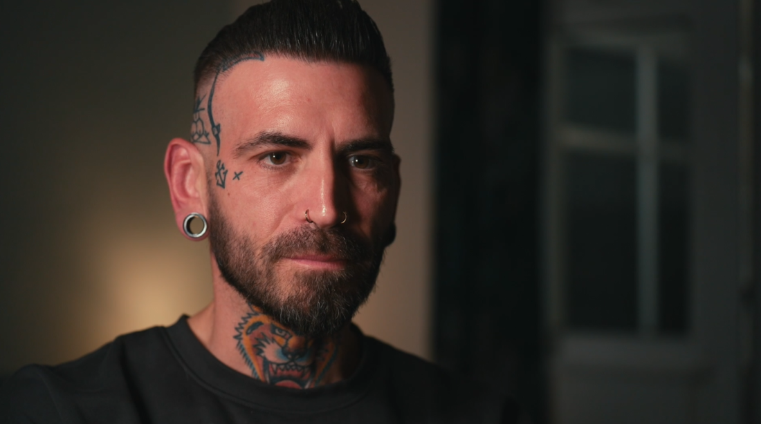 CURBB participant Sam is seated facing the camera. He is wearing a black shirt. The image is cropped to head and shoulders. He has short brown hair, a beard, numerous piercings and tattoos.